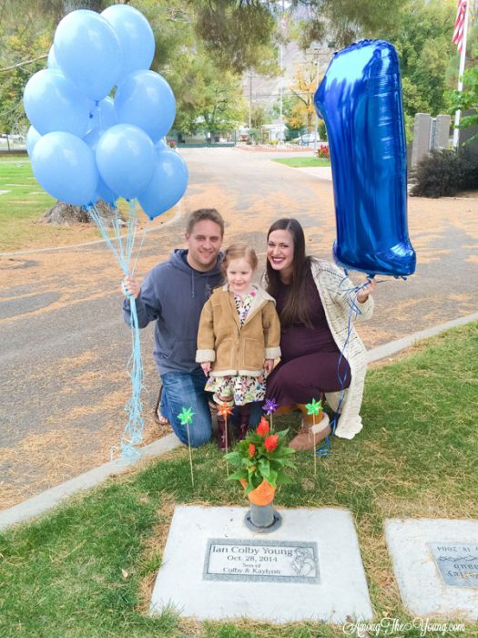 sibling loss featured by top Utah lifestyle blog, Among the Young: image of first birthday | Sibling Loss: 7 Essential Tips to Help your Child Grieve by popular Utah lifestyle blog, Among the Young: image of a family kneeling down in front of a headstone and holding blue balloons.