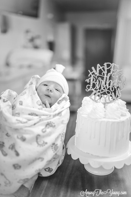 Baby girl birth story by top Utah lifestyle blog, Among the Young: image of baby and birthday cake | Birth Story by popular Utah motherhood blog, Among the Young: black and white image of a newborn baby girl being held next to a layer cake with a Happy Birthday cake topper. 