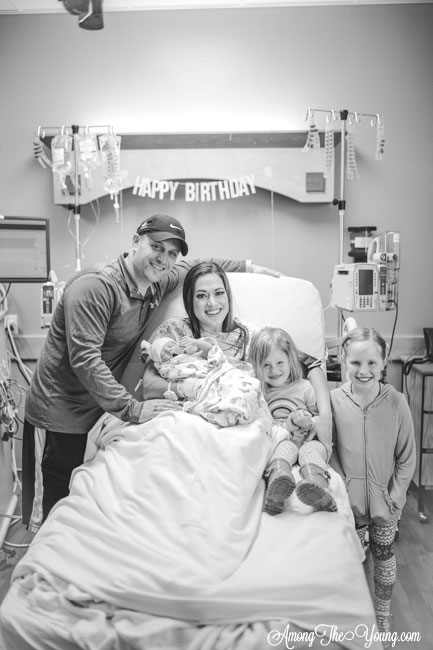 Baby girl birth story by top Utah lifestyle blog, Among the Young: image of new family of 6 | Birth Story by popular Utah motherhood blog, Among the Young: black and white image a mom sitting in a hospital bed and holding her newborn daughter in her arms while she is surrounded by her husband and two other daughters. 